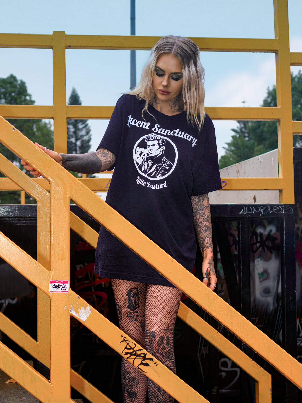 A woman with blonde hair stands on yellow metal stairs wearing a black James Dean graphic t-shirt with 'Lucent Sanctuary' and  printed on the front, paired with fishnet stockings.